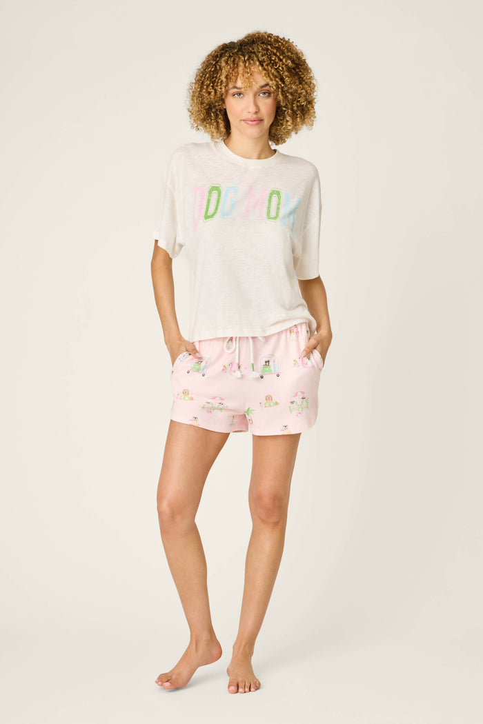 A woman with curly hair stands barefoot against a light background, wearing P.J. Salvage’s Pupcation Palms Sunnies & Sunshine PJ Shorts in peach and a loose white T-shirt with colorful text, hands in her pockets.