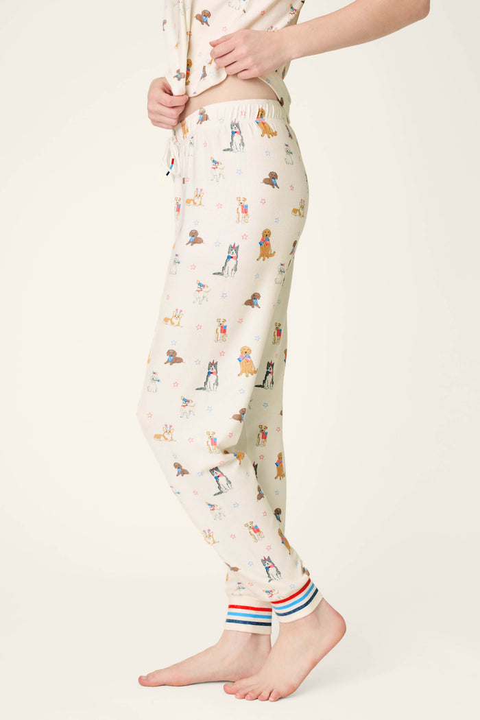 A person in P.J. Salvage Red, White, & Woof Patriotic Pups Banded Pants with multi-colored striped cuffs stands barefoot on a light background. The playful dog print and soft fabric offer cozy comfort for relaxed days at home. Upper body partially visible.