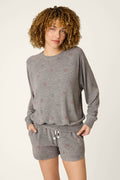 A person with curly hair wears the P.J. Salvage Red, White, & Woof Star Print Long Sleeve Top—a relaxed fit gray lounge top with small red and blue stars—paired with matching drawstring shorts, hands in pockets against a plain background.
