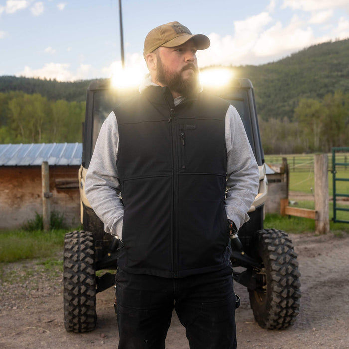 Man wearing a black Rexford Ripstop vest over a gray hoodie, standing in front of an off-road vehicle in a rural setting.