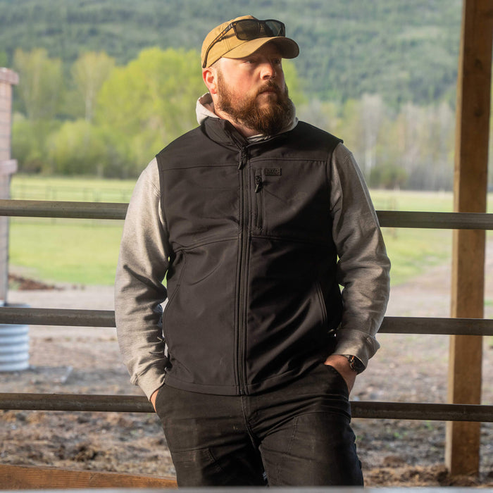 Man wearing a black Rexford Ripstop Vest over a gray hoodie, standing outdoors in a rural setting.