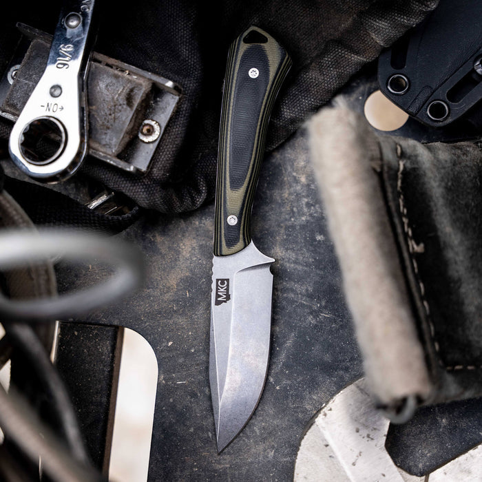 Rocker knife in green and black, featuring a sleek blade and ergonomic handle, resting on a rugged surface.