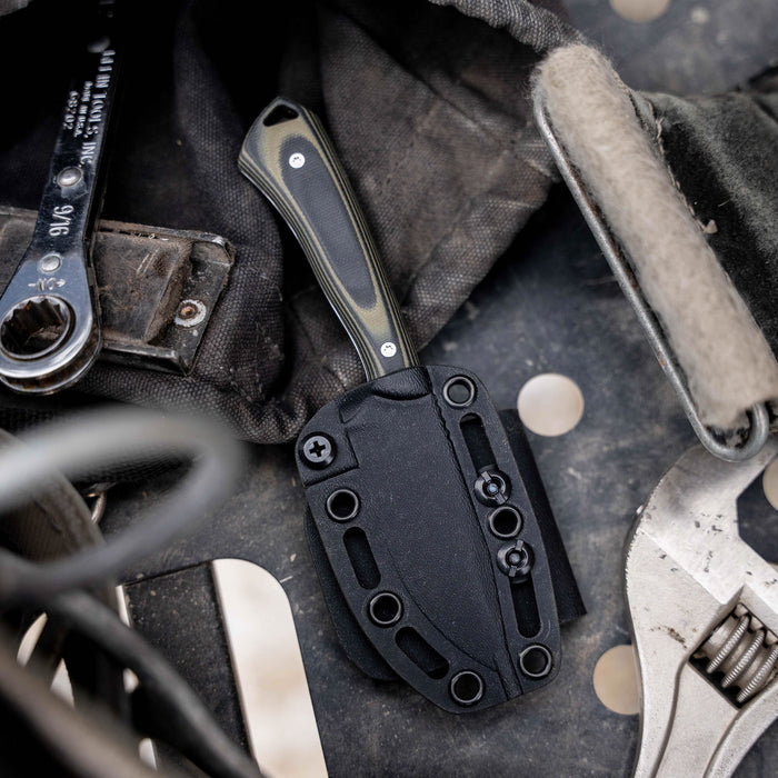 Green and black ROCKER knife in a black sheath, resting on a textured surface with tools and gear nearby.