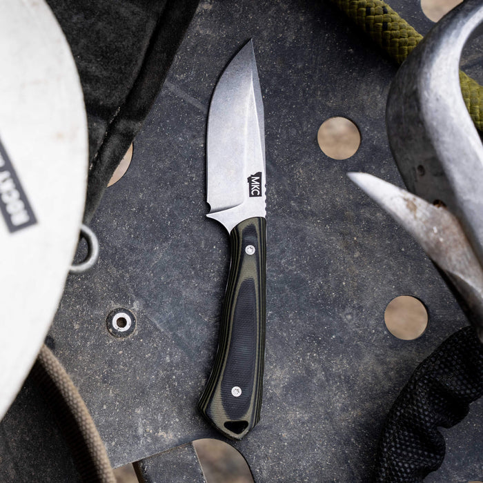 Rocker knife in green and black with a durable handle, resting on a rugged surface among outdoor gear.