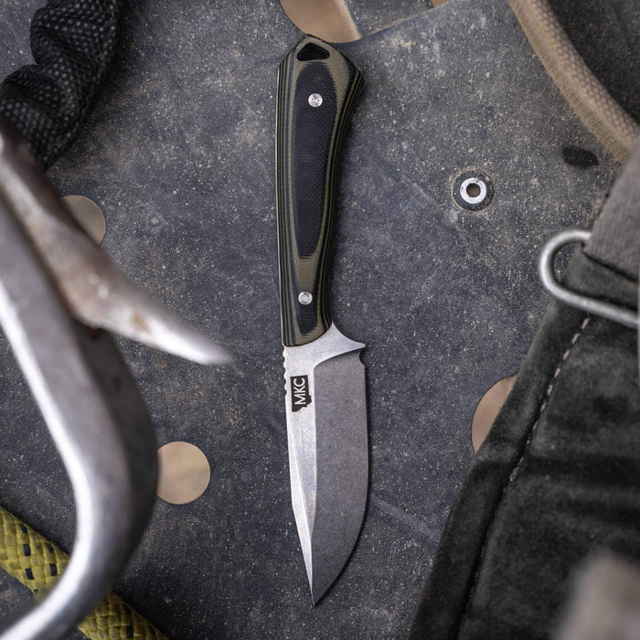 Rocker knife in green and black with a durable blade, resting on textured surface with climbing gear.