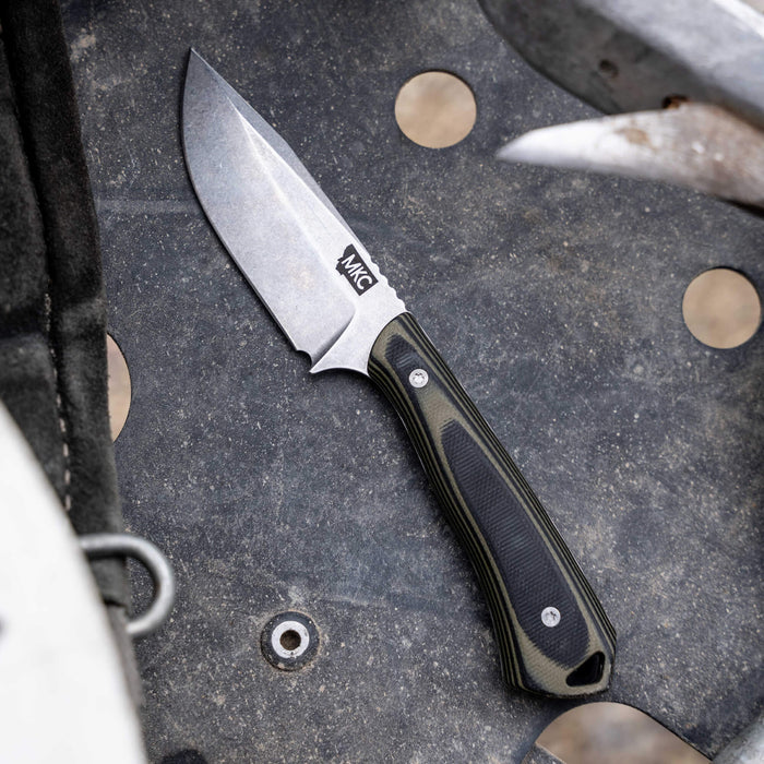 ROCKER knife in green and black, featuring a rugged blade and textured handle, resting on a metal surface.