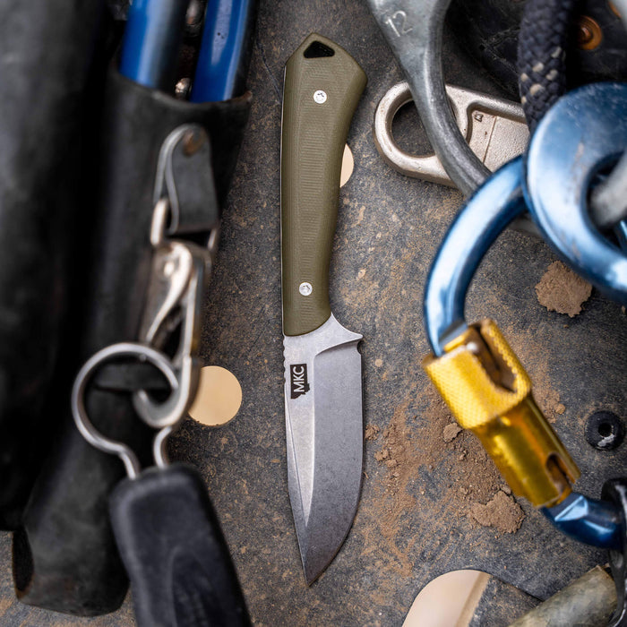 Olive green ROCKER knife resting on a rugged surface among climbing gear and tools, showcasing its durable design.