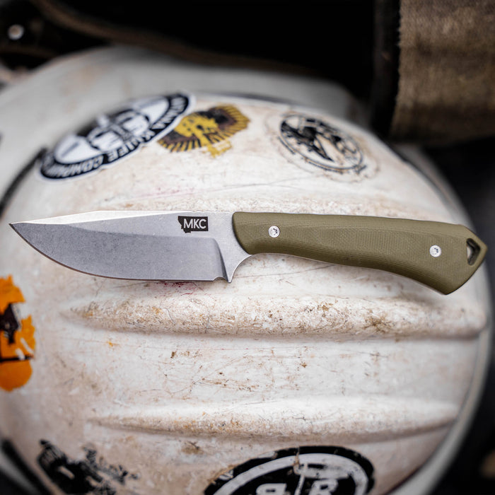 Olive-handled ROCKER knife by Montana Knife Company on a textured surface, showcasing its durable blade and ergonomic design.
