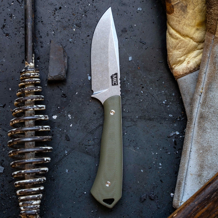 Olive green fixed-blade knife with a curved blade, set on a textured surface alongside tools and leather.