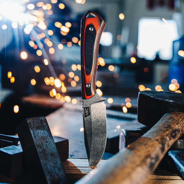 ROCKER knife in orange and black, displayed among tools with sparks flying, showcasing its rugged design and craftsmanship.