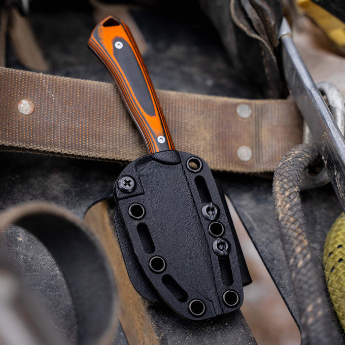 ROCKER knife with an orange and black handle in a black sheath, resting on a tool belt in an outdoor setting.