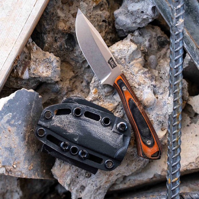 Rocker knife in orange and black with a textured handle, displayed on rubble alongside its durable sheath.