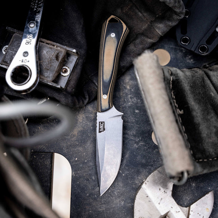 Tan and black ROCKER knife with a sleek blade, resting on a rugged surface among tools and gear.