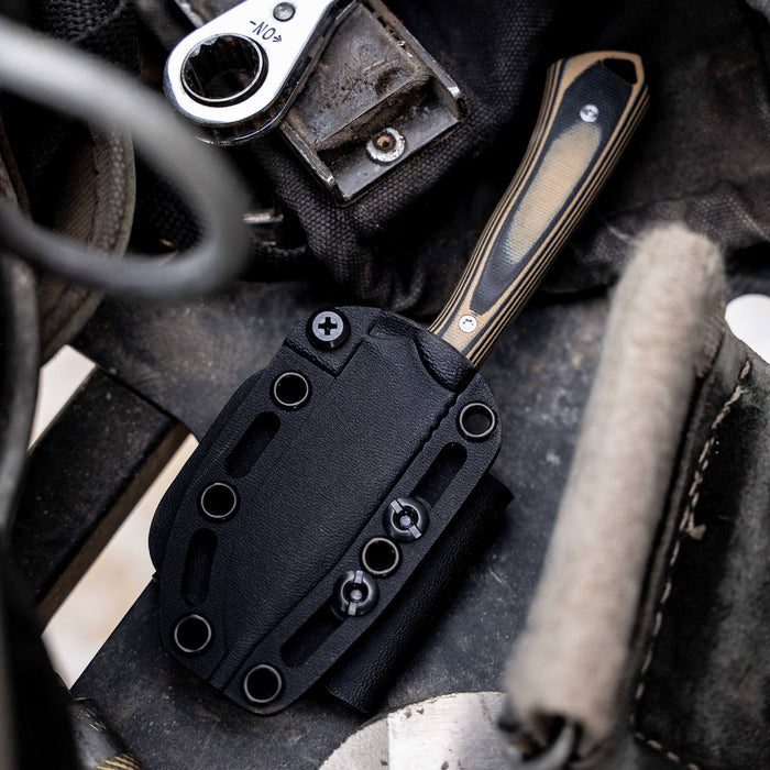 Rocker knife in tan and black sheath, resting on a rugged surface with tools, showcasing its durable design for outdoor use.