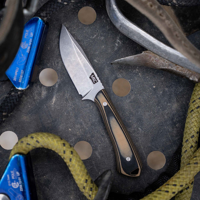 Rocker knife in tan and black, featuring a fixed blade, resting on a textured surface with climbing gear.