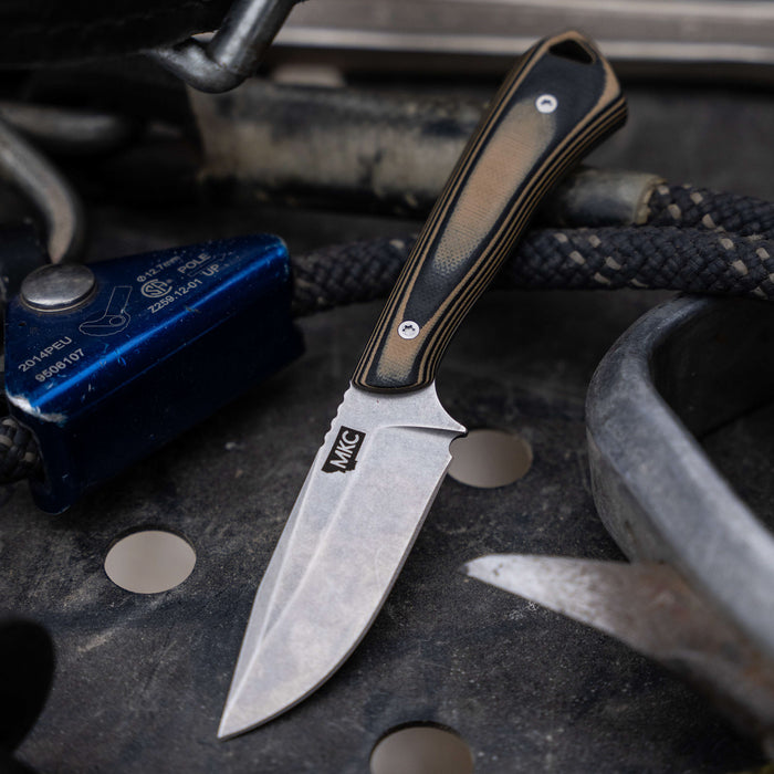 Rocker knife in tan and black with a textured handle, resting on a rugged surface among tools and equipment.