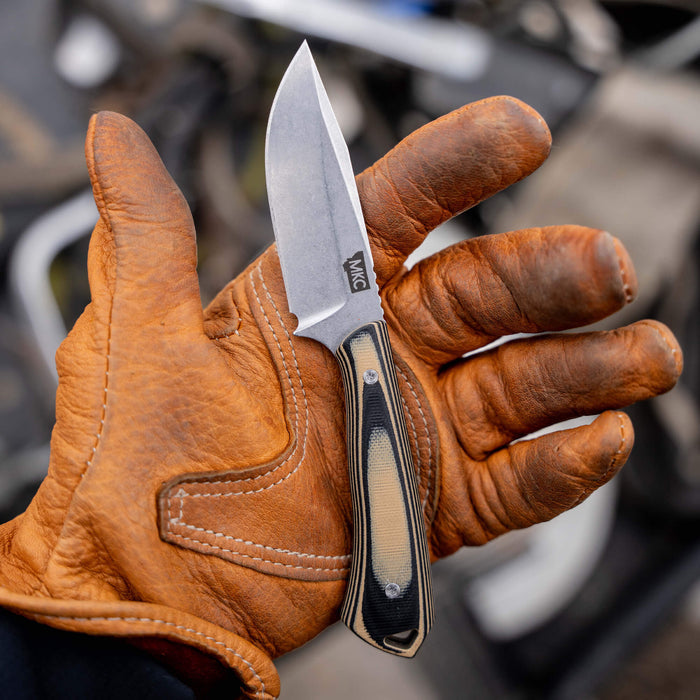 A tan and black ROCKER knife held in a gloved hand, showcasing its sleek design and durable blade, ideal for outdoor use.