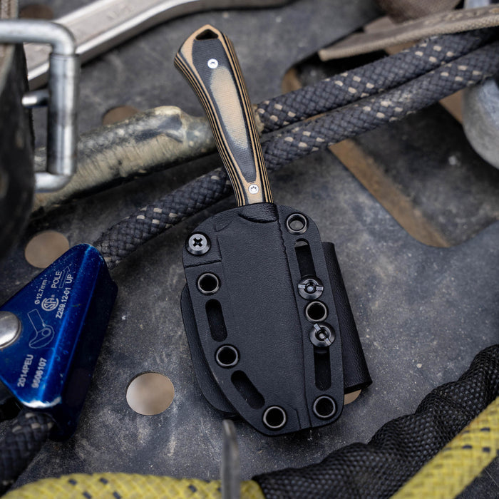 Rocker knife in tan and black with a textured handle, secured in a black sheath on a rugged surface with gear.