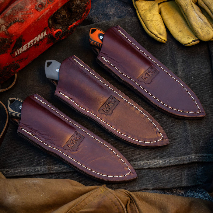 Three vertical leather sheaths for knives, featuring sturdy stitching and the MKC logo, placed on a rugged work surface.