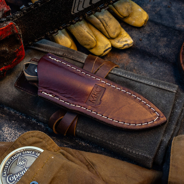 Vertical leather sheath for knives, featuring durable construction and MKC branding, set against outdoor tools and gloves.
