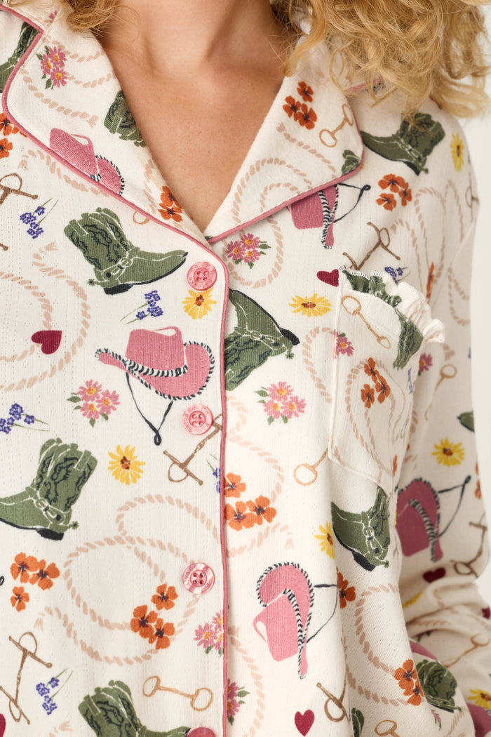 Close-up of the Saddle Up Western Print Long Sleeve PJ Set by P.J. Salvage, showcasing the pajama top’s cream background with pink hats, green boots, flowers, horseshoes, hearts, and lassos in a Western-themed print. The top features pink piping and visible buttons.