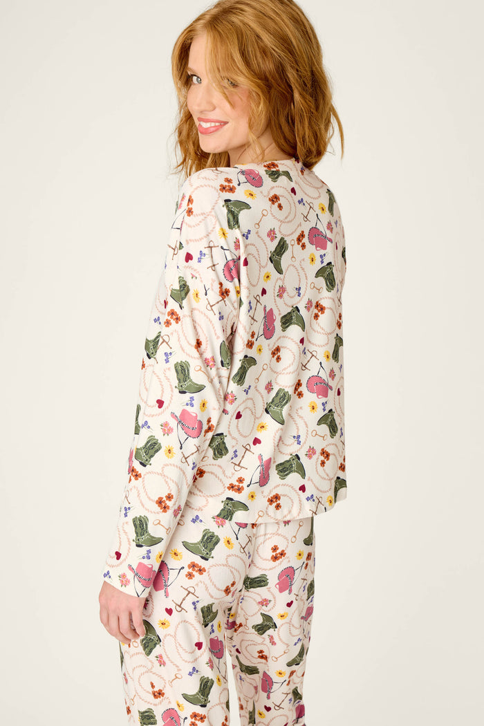A woman with red hair smiles while looking back over her shoulder. She is wearing the P.J. Salvage Saddle Up Western Print Long Sleeve V-Neck Top, which features playful patterns and images on a white background.
