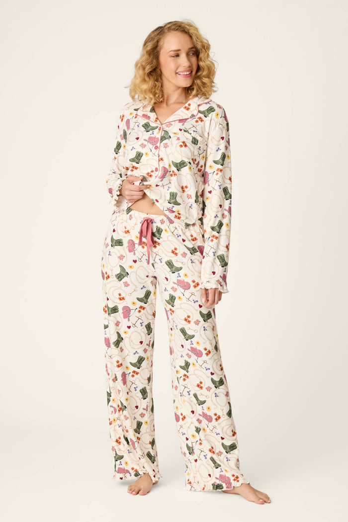 A woman with curly blonde hair stands barefoot and smiles against a plain light background, wearing the P.J. Salvage Saddle Up Western Print Pajama Pants. The pants feature a long, relaxed fit with button-up detailing, floral prints accented by pink trim and a pink drawstring at the waist.