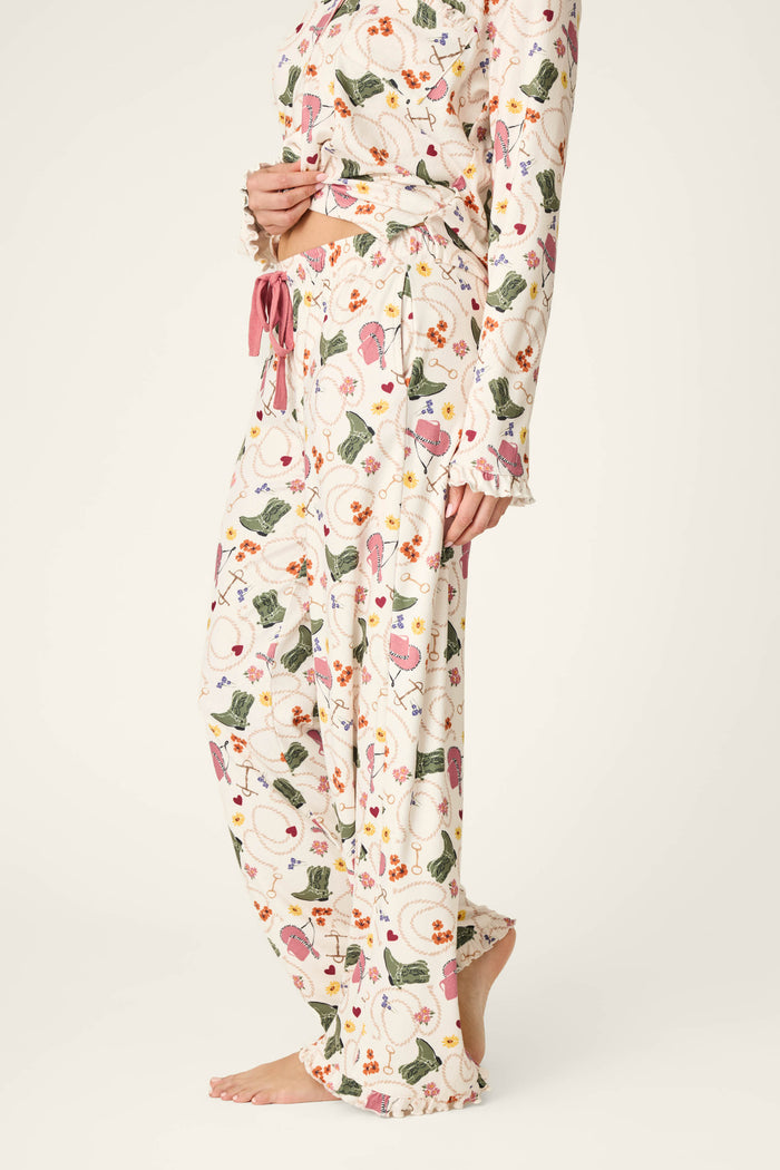 A person standing barefoot on a neutral background is shown from the lower body and midsection, wearing P.J. Salvage Saddle Up Western Print Pajama Pants featuring a colorful floral and animal pattern.