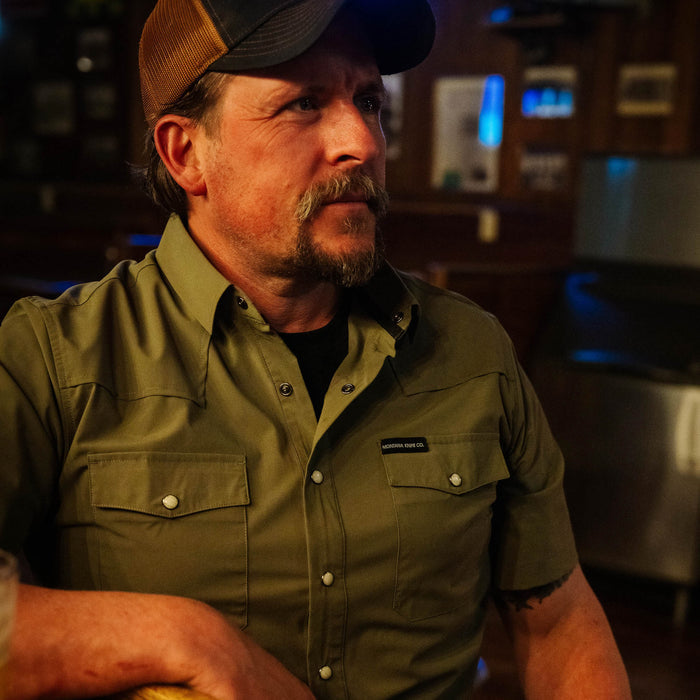 Olive short-sleeve button-up shirt worn by a man in a rustic setting, showcasing a casual yet rugged style.