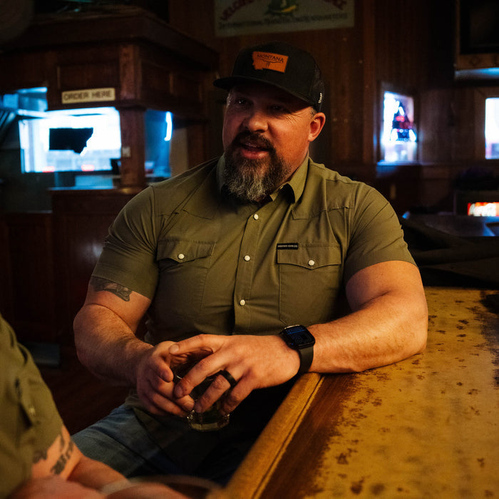 Man wearing an olive short-sleeve button-up shirt, seated at a bar, holding a drink, showcasing a rugged style.