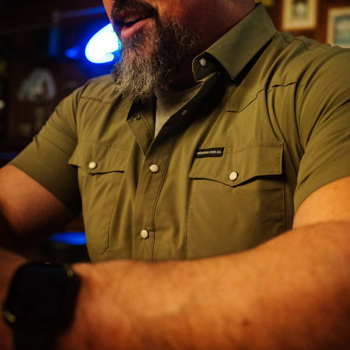 Olive short-sleeve button-up shirt worn by a man, featuring two chest pockets and a Montana Knife Company logo.