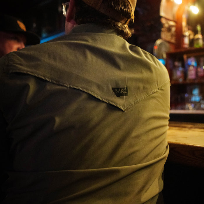 Olive short-sleeve button-up shirt from Montana Knife Company, featuring the MKC logo on the back, worn in a casual setting.