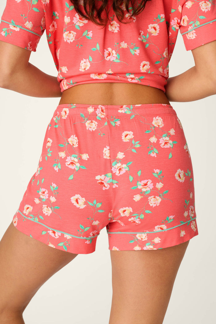 A woman is seen from the back wearing P.J. Salvage's Siesta Dreams Dark Coral Floral Print Sleep Shorts, a modal jersey pajama set with short sleeves and shorts featuring vibrant flowers and green leaves.