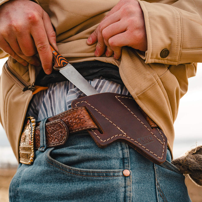 A person wearing a beige jacket and denim jeans retrieves the STOCKYARD knife from a leather sheath on their belt.
