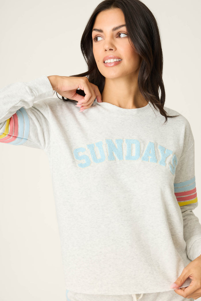 A woman wearing the P.J. Salvage Sunday Funday Boucle "Sundays" Sweatshirt, featuring blue lettering and pastel rainbow sleeve stripes, looks to the side and smiles with one hand touching her collar.