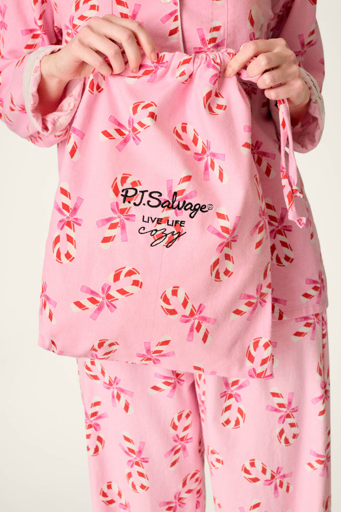 A person wearing the P.J. Salvage Sweeter Than Sugar Candy Cane Print 4-Piece PJ Set, featuring pink pajamas with red and white candy cane patterns, holds a matching drawstring bag that says “P.J. Salvage LIVE LIFE cozy” in black text.