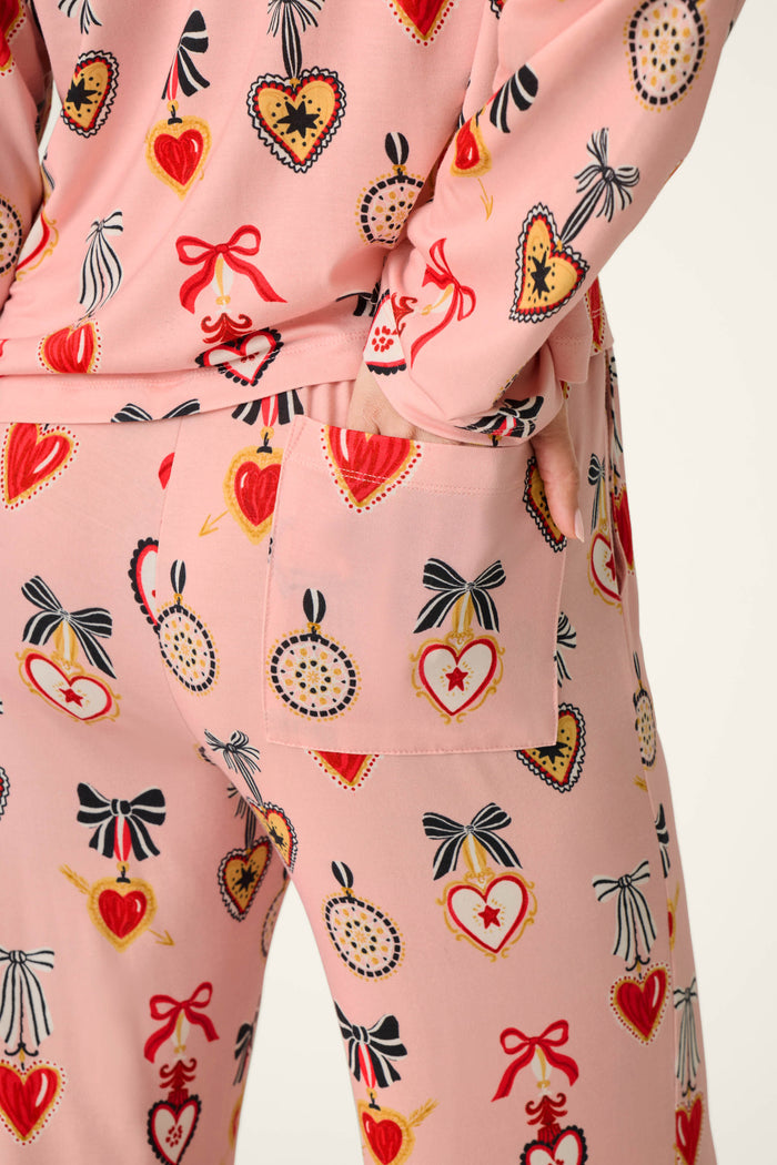 A person is wearing the P.J. Salvage Sweetheart Dreams Love Lockets Print Long Sleeve PJ Set, featuring red and yellow heart and bow motifs. They have one hand placed in the pocket of the pants, standing against a plain, off-white background.
