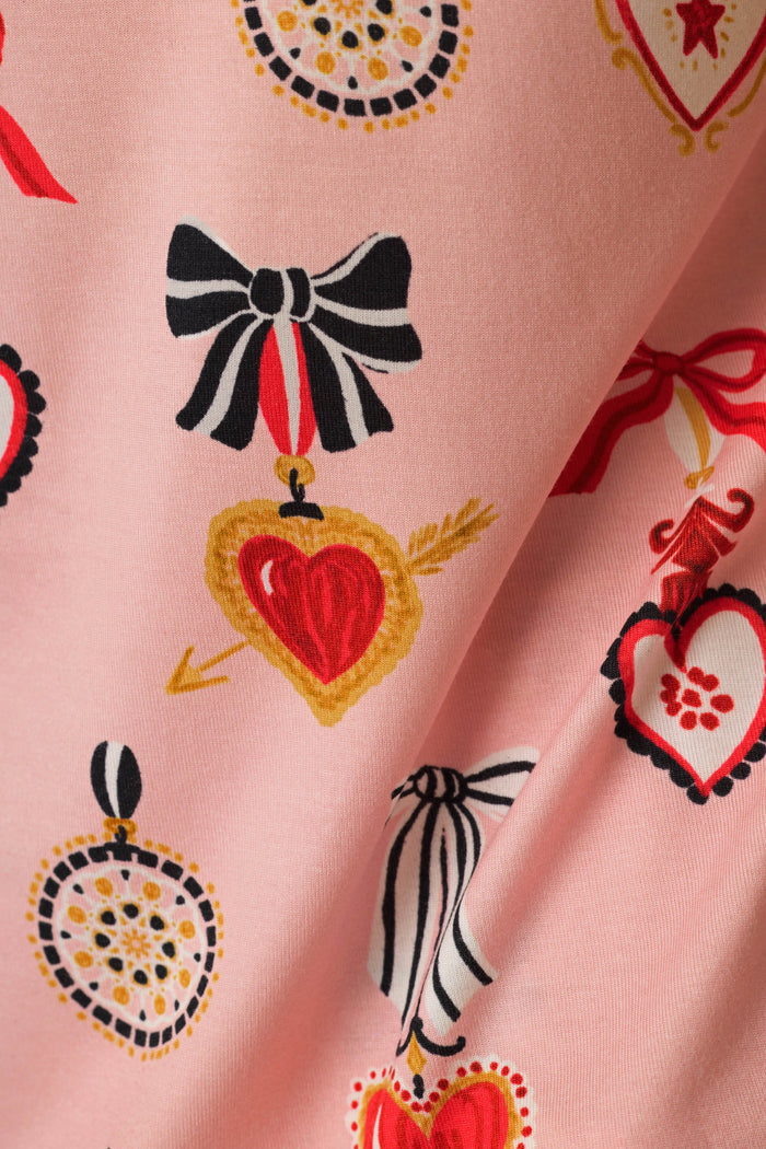 A close-up of a light pink fabric with a repeating pattern of stylized red hearts, black-and-white bows, and ornate gold and black decorative motifs.