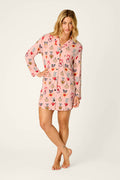 A woman stands barefoot wearing the P.J. Salvage Sweetheart Dreams Love Lockets Print Nightshirt, a pink, long-sleeved nightshirt adorned with colorful love locket prints. She smiles slightly, placing one hand on her hip and bending one leg gently.