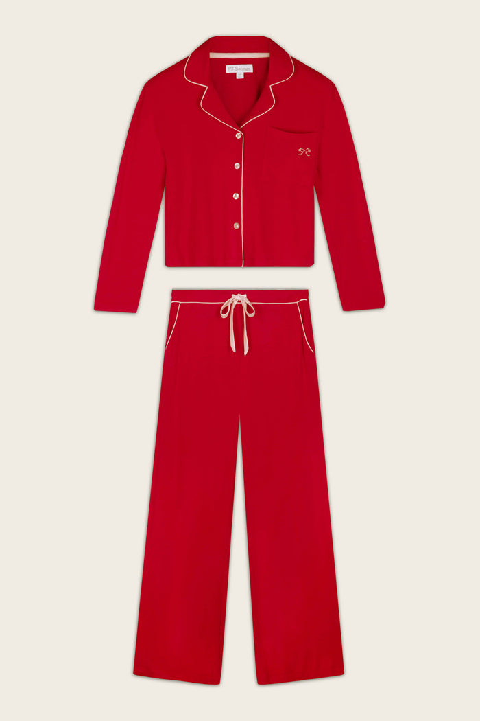 A two-piece red pajama set with cream-colored piping and trim, consisting of a long-sleeve button-front top with a collar, that has a bow embroidered on the chest pocket, and matching drawstring pants, displayed flat against a light beige background.