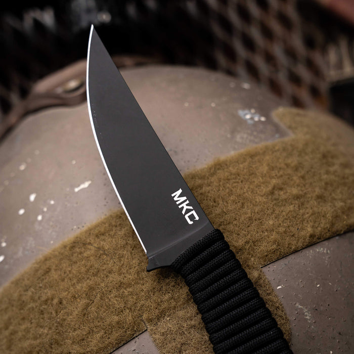 Tactical Speedgoat knife with a black blade and paracord handle, resting on a textured surface.