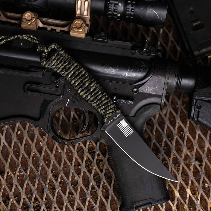 Tactical Speedgoat knife with black and dark camo handle, positioned next to a firearm on a metal surface.