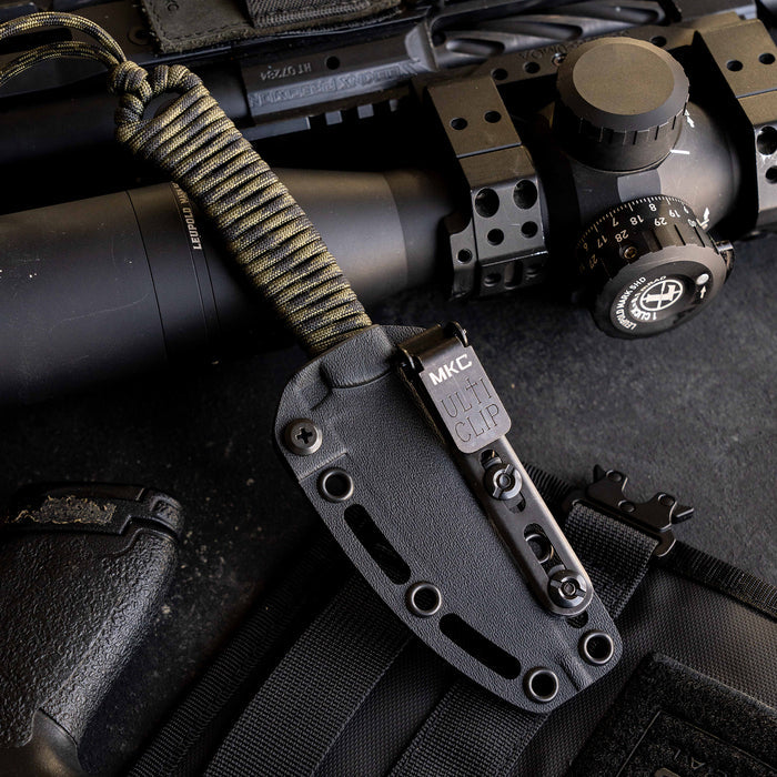 Kydex sheath for Tactical Speedgoat knife, featuring a utility clip, positioned near tactical gear and a rifle scope.