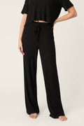 A person wearing P.J. Salvage Textured Essentials Black Open Leg Pants with a drawstring waistband and a black short-sleeve top stands barefoot against a plain light background.