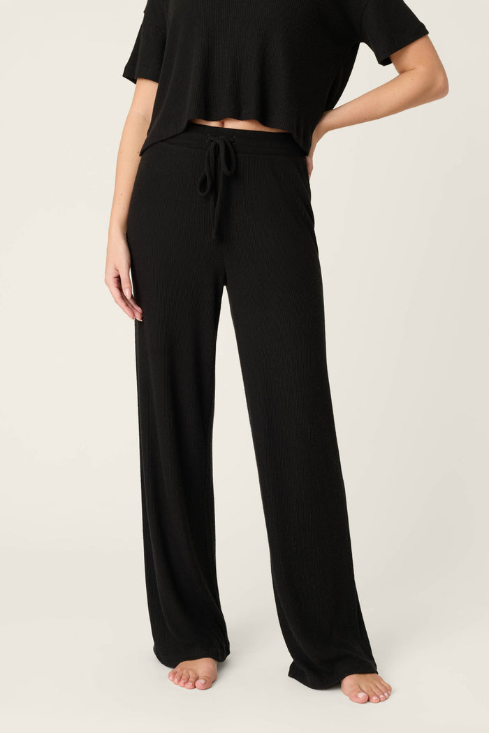 A person wearing P.J. Salvage Textured Essentials Black Open Leg Pants with a drawstring waistband and a black short-sleeve top stands barefoot against a plain light background.