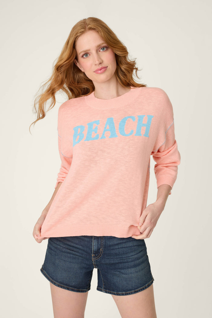 A person with long, wavy hair wears a light pink sweater with "BEACH" in blue letters and blue denim shorts on a plain background.