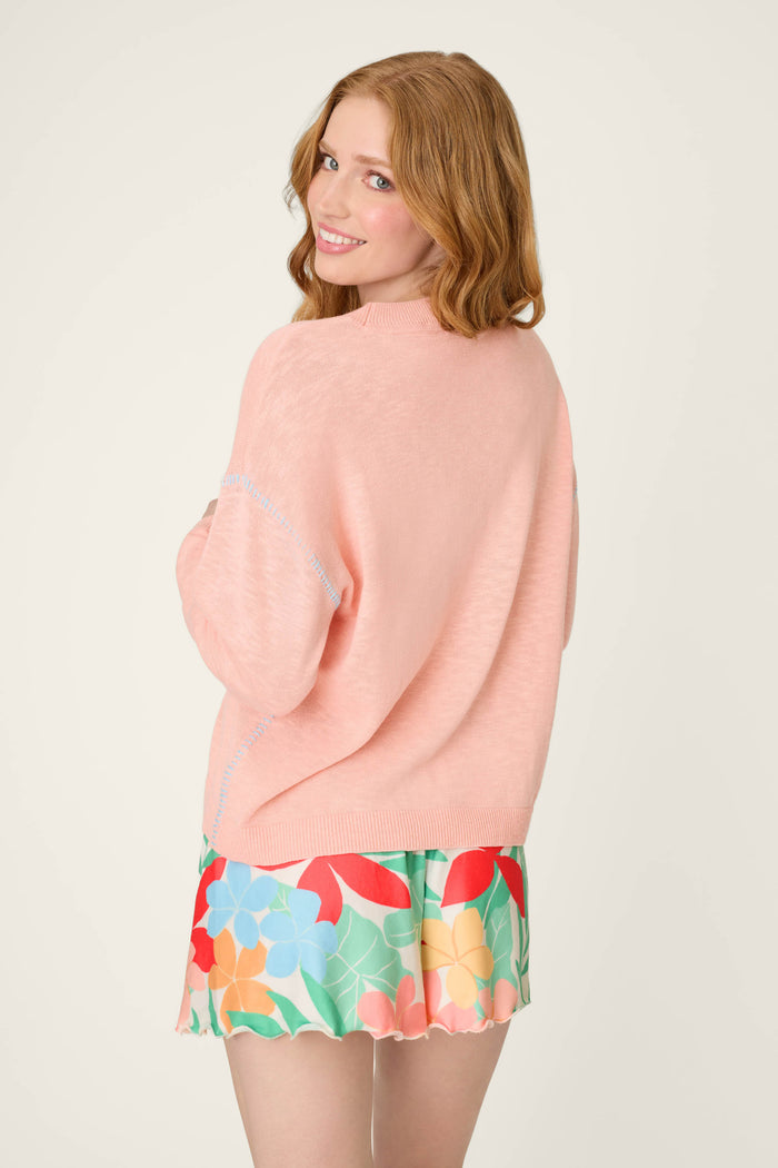A person with long, light brown hair stands against an off-white background wearing a lightweight pink sweater.