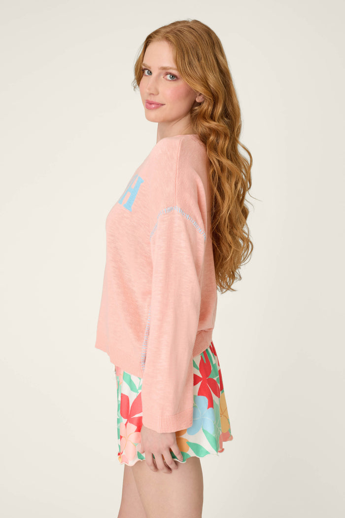 A woman with long wavy red hair stands sideways, looking at the camera in a beach sweater and colorful floral shorts on a plain background.