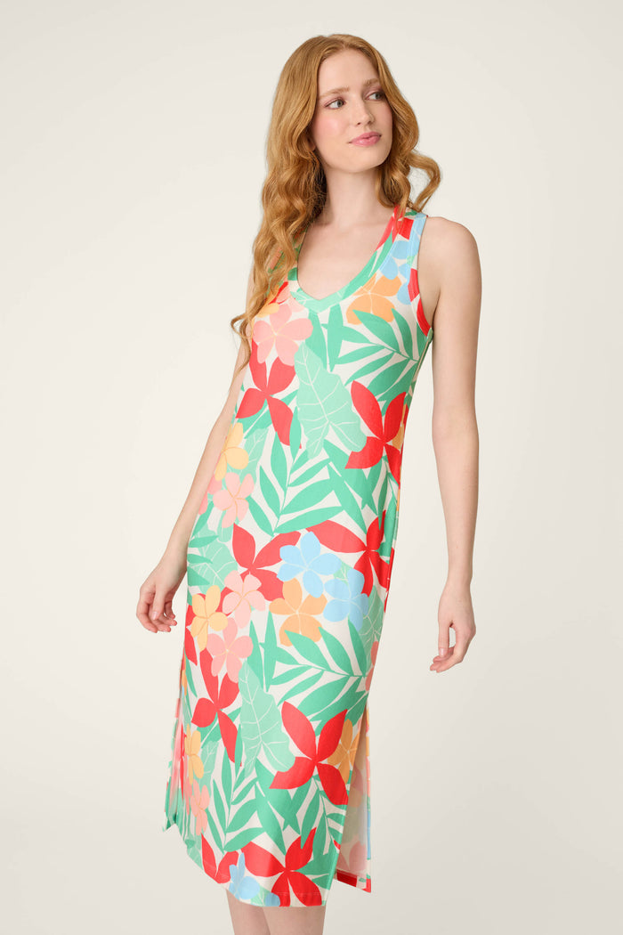 A woman with long, wavy red hair wears a sleeveless, knee-length lounge dress in a colorful tropical print, standing against a plain light background and looking slightly to the side.