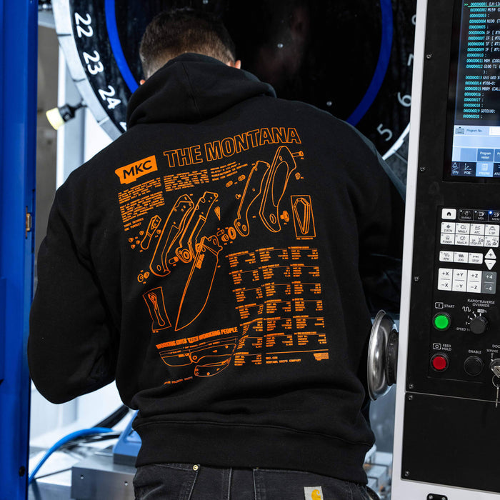Black Montana Hoodie featuring orange graphic design showcasing knife specifications, worn by a person in a workshop.